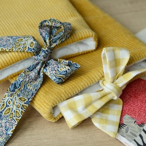 May include: Two rectangular mustard yellow corduroy sleeves with white interior lining. One sleeve features a blue and multi-colored floral patterned fabric tie, while the other features a yellow and white gingham patterned fabric tie.