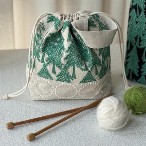 May include: A drawstring bag with a green tree pattern, a beige bottom, and a small pocket. Two wooden knitting needles, a ball of white yarn, and a ball of green yarn are in front of the bag.