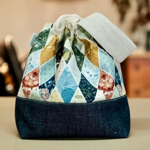 Könnte beinhalten: Ein Kordelzugbeutel mit einem Patchwork-Design. Der obere Teil hat ein Rautenmuster in Blau-, Grün-, Gelb- und Rottönen. Der Boden ist aus dunkelblauem Denim. Oben befinden sich eine weiße Kordel und ein graues Stoffstück.