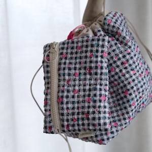 May include: A set of two fabric bags, one rectangular with a zipper and the other a drawstring bag. Both are patterned with a black and white gingham check and small pink and blue floral accents. The bags are hanging from a neutral-colored cord.