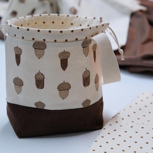 May include: A cylindrical fabric drawstring bag featuring a cream-colored upper section with a brown acorn print and a brown polka dot trim. The lower portion is made of dark brown corduroy fabric. It includes a side handle and drawstring closure.