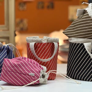 May include: Several striped fabric bags in navy, pink, red, black, and brown. Each bag has white stripes, a drawstring closure, and neutral-toned trim. Some bags have handles. The bags are for storage.
