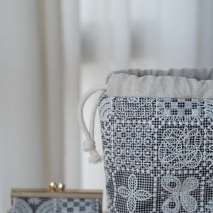 Peut inclure: Une pochette &agrave; cordon dot&eacute;e d'une base en tissu pied-de-poule noir et blanc avec des motifs g&eacute;om&eacute;triques et floraux blancs brod&eacute;s. La partie sup&eacute;rieure est en tissu beige uni avec un cordon de serrage blanc &eacute;pais.