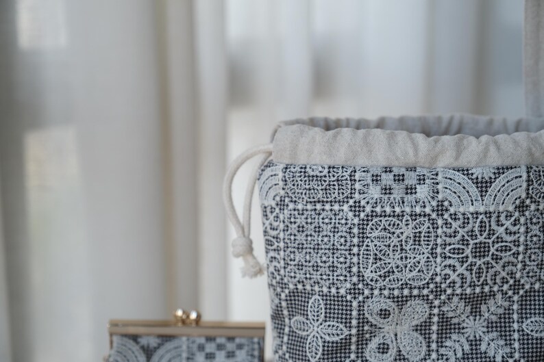 Peut inclure: Une pochette &agrave; cordon dot&eacute;e d'une base en tissu pied-de-poule noir et blanc avec des motifs g&eacute;om&eacute;triques et floraux blancs brod&eacute;s. La partie sup&eacute;rieure est en tissu beige uni avec un cordon de serrage blanc &eacute;pais.