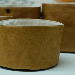 Könnte beinhalten: Ein Set aus drei braunen Aufbewahrungsbehältern mit strukturierter Oberfläche. Die Behälter haben eine zylindrische Form mit weißem Innenfutter. Sie bestehen aus einem strapazierfähigen Material und eignen sich zum Ordnen verschiedener Gegenstände.