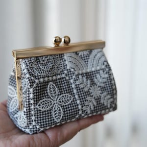 Peut inclure: Un petit porte-monnaie en tissu &agrave; motif pied-de-poule noir et blanc, orn&eacute; de broderies florales et v&eacute;g&eacute;tales blanches. L'accessoire est dot&eacute; d'une fermeture &agrave; fermoir m&eacute;tallique dor&eacute; de type porte-monnaie &agrave; l'ouverture.