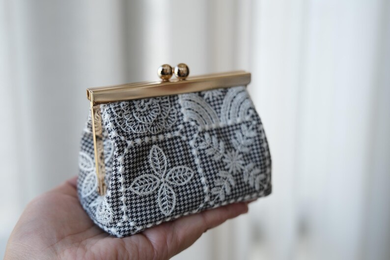 Peut inclure: Un petit porte-monnaie en tissu &agrave; motif pied-de-poule noir et blanc, orn&eacute; de broderies florales et v&eacute;g&eacute;tales blanches. L'accessoire est dot&eacute; d'une fermeture &agrave; fermoir m&eacute;tallique dor&eacute; de type porte-monnaie &agrave; l'ouverture.