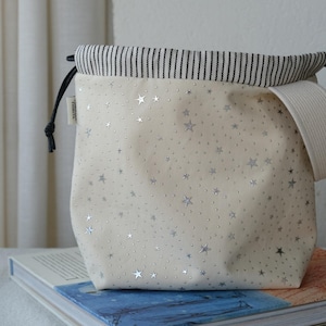 Può includere: Borsa a coulisse color crema con un motivo a stelle argentate. Il bordo superiore ha un tessuto a righe bianche e nere e un manico color crema è attaccato al lato. I cordini neri chiudono la borsa.