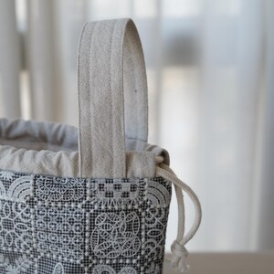 Peut inclure: Une pochette &agrave; cordon dot&eacute;e d'un motif en dentelle g&eacute;om&eacute;trique noir et blanc. Le sac pr&eacute;sente une partie sup&eacute;rieure en tissu beige avec une anse assortie et une fermeture &agrave; cordon blanc. L'ext&eacute;rieur affiche un motif &agrave; grille carr&eacute;e r&eacute;p&eacute;titif.