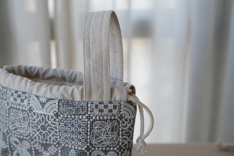 Peut inclure: Une pochette &agrave; cordon dot&eacute;e d'un motif en dentelle g&eacute;om&eacute;trique noir et blanc. Le sac pr&eacute;sente une partie sup&eacute;rieure en tissu beige avec une anse assortie et une fermeture &agrave; cordon blanc. L'ext&eacute;rieur affiche un motif &agrave; grille carr&eacute;e r&eacute;p&eacute;titif.