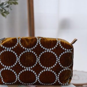 May include: A rectangular mustard-colored velvet pouch featuring a repeating pattern of white embroidered circles. The bag has a top zipper closure with small fabric tabs on each side and a small white brand tag attached to the left side seam.