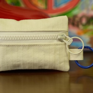 May include: A small, rectangular off-white pouch with a white zipper and a blue ring. The pouch has a textured surface and a small loop with a white ring attached to a blue ring. The background is blurred.