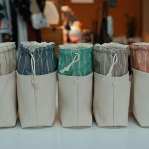 Puede incluir: Cinco bolsas de almacenamiento de lona con la parte superior con cordón. Las bolsas están en fila, cada una con una parte superior de tela a rayas diferente en tonos beige, azul, verde, gris y coral. Las bolsas son de color blanquecino y tienen un diseño simple y minimalista.