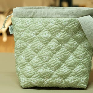 May include: A light green quilted fabric drawstring bag with a diamond pattern. The top edge and a small strap are a lighter green. A small tag is attached to the side. The bag is sitting on a light brown surface.