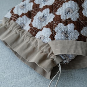 May include: A quilted bonnet featuring a floral pattern in white and brown. The bonnet has a beige drawstring and a ruffled edge. The fabric is soft and the design is detailed, suggesting a handmade item.