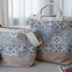 May include: Two drawstring bags featuring a dark gray and white patchwork lace pattern on the upper section and a solid beige corduroy base. The bags have light beige fabric handles and drawstring closures, with a ball of blue yarn nearby.