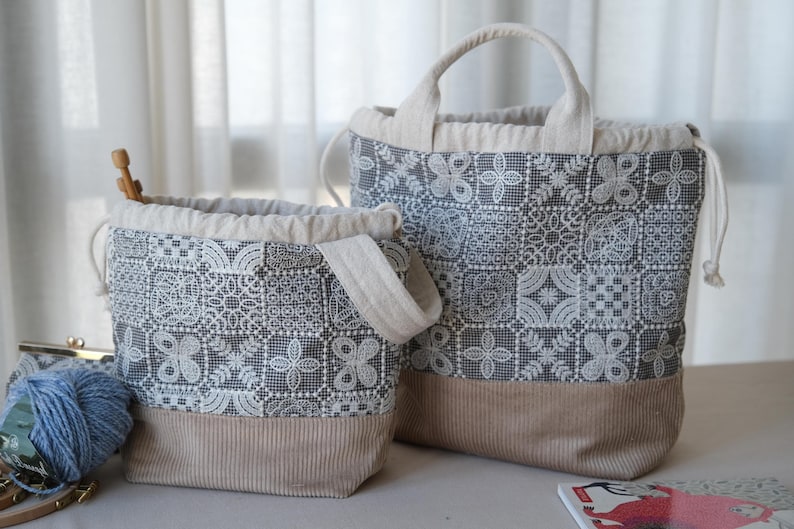 Peut inclure: Deux sacs &agrave; cordon dot&eacute;s d'un motif en dentelle patchwork gris fonc&eacute; et blanc sur la partie sup&eacute;rieure et d'une base en velours c&ocirc;tel&eacute; beige uni. Les sacs ont des anses en tissu beige clair et des fermetures &agrave; cordon, avec une pelote de laine bleue.