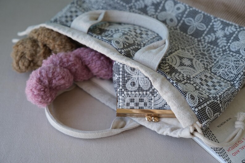 Peut inclure: Un sac fourre-tout en tissu avec un motif de dentelle g&eacute;om&eacute;trique noir et blanc et deux anses. &Agrave; l'int&eacute;rieur se trouvent deux &eacute;cheveaux de laine textur&eacute;e marron et rose, ainsi qu'un petit porte-monnaie &agrave; cadre m&eacute;tallique et fermoir dor&eacute;.