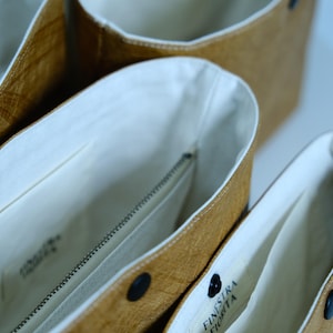 Op de afbeelding: Close-up van drie open tote tassen. De tassen hebben een lichtbruine buitenkant en een witte binnenkant. Elke tas heeft een zwarte drukknoopsluiting. De binnenkant van de tassen heeft een rits en een label met tekst. De tassen zijn gemaakt van een textuurrijk materiaal.