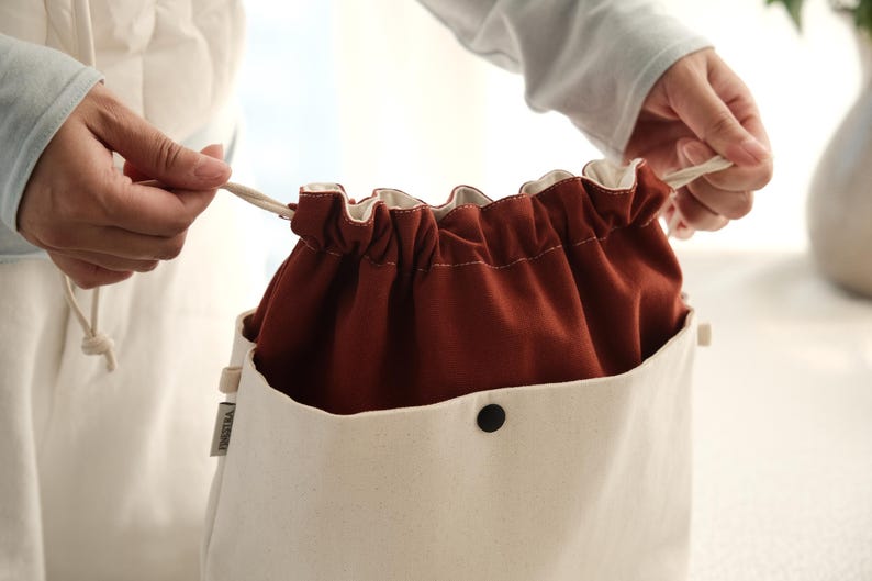 May include: A two-toned drawstring bag featuring a cream-colored exterior pocket with a black snap button closure and a deep rust-colored fabric top section. The bag includes a cream drawstring cord and a small side tag with visible text.