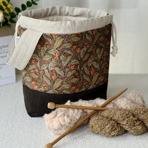 May include: A cylindrical knitting project bag with a floral pattern in brown, green, and orange. The bag has a dark brown base and a beige drawstring top. Two wooden knitting needles and yarn are in front of the bag.