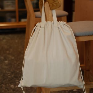 Può includere: Una borsa beige con coulisse e manici abbinati è appesa a una sedia di legno. La borsa è realizzata in un tessuto morbido e chiaro e ha un design semplice e minimalista. La borsa è chiusa con un cordoncino.
