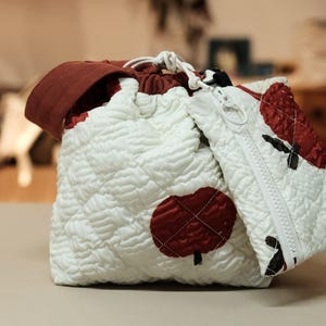 May include: A white quilted drawstring bag and matching zippered pouch with red apple and black leaf designs. The bag has a burgundy rim and a white drawstring closure. The pouch has a white zipper.