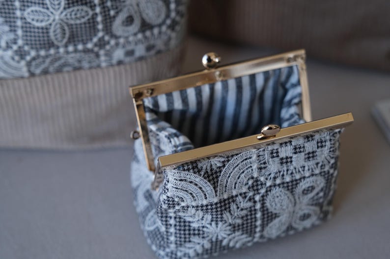 Peut inclure: Un petit porte-monnaie en tissu pied-de-poule noir et blanc avec une broderie en dentelle florale blanche. Il est dot&eacute; d'un fermoir &agrave; cliquet en m&eacute;tal dor&eacute; et d'une doublure int&eacute;rieure en tissu ray&eacute; noir et blanc.