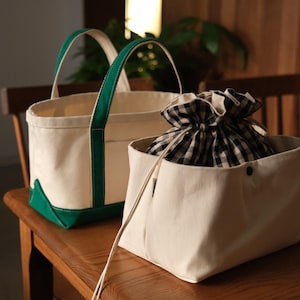 Puede incluir: Dos bolsas de lona. Una es de color crema con asas y base verdes. La otra es una bolsa rectangular de color crema con cierre de cordón y una tela a cuadros en blanco y negro en el interior.