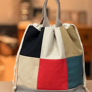 May include: A patchwork drawstring bag with a variety of colors including black, red, beige, and teal. The bag has a gray base and a beige handle. The drawstring is white.