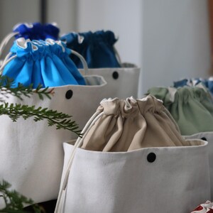 May include: A collection of white canvas bucket bags featuring drawstring closures in various solid colors including bright blue, dark teal, beige, and sage green. Each bag has a single black snap button on the front and white cord drawstrings.