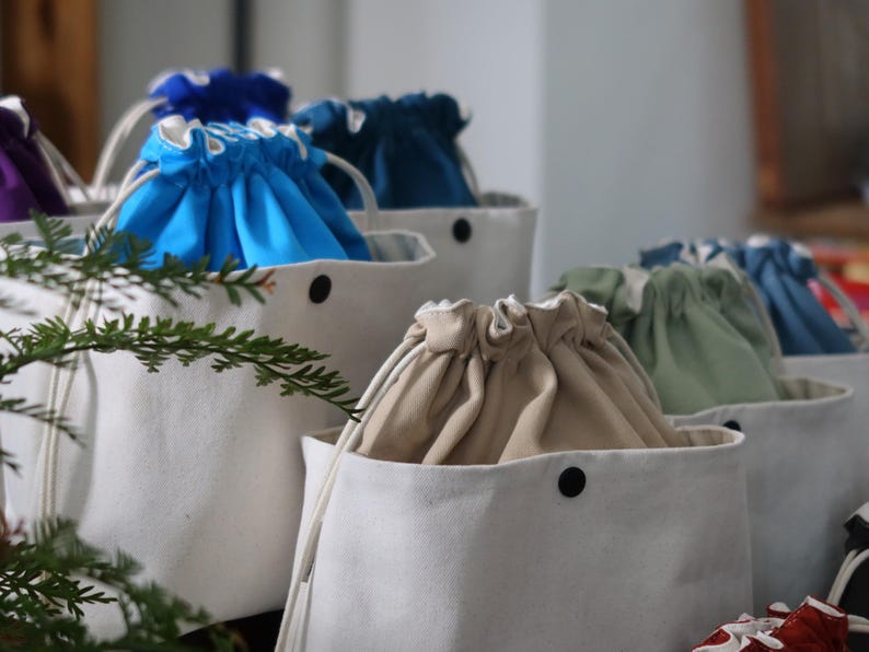 May include: A collection of white canvas bucket bags featuring drawstring closures in various solid colors including bright blue, dark teal, beige, and sage green. Each bag has a single black snap button on the front and white cord drawstrings.
