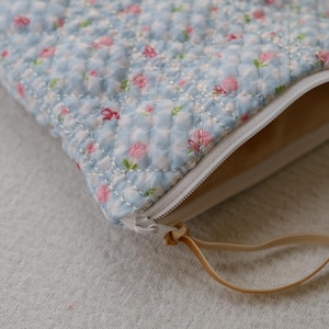 Könnte beinhalten: Ein gesteppter Beutel mit einem hellblauen Blumenmuster mit rosa Blüten und grünen Blättern. Der Beutel hat einen weißen Reißverschluss und ein beiges Innenfutter. Eine beige Schlaufe ist am Reißverschluss befestigt.