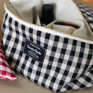 Puede incluir: Bolsa con cremallera de cuadros blancos y negros con cremallera dorada, que contiene una botella negra. Una almohada de cuadros rojos y blancos está en el fondo. La bolsa tiene una etiqueta que dice "Finestra Fiorita, Crafted with Care."