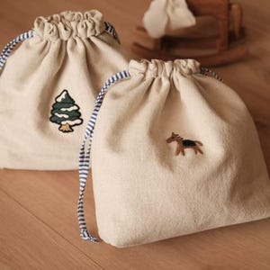 May include: Two beige drawstring bags with embroidered designs. One features a green and white tree, the other a brown and black horse. The bags have blue and white striped drawstrings and are made of a soft, textured fabric.