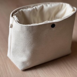 May include: An off-white canvas pouch with a structured rectangular base and a soft, gathered opening. The bag features a single black snap button closure centered at the top edge and a small fabric loop on the side for attachment or carrying.