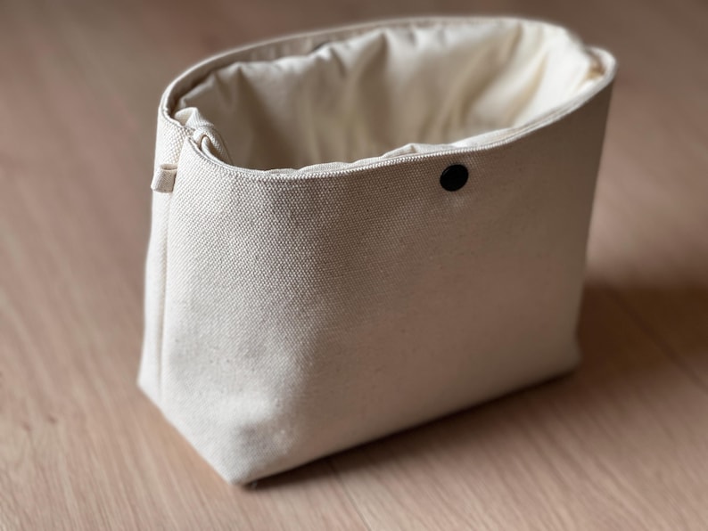 May include: An off-white canvas pouch with a structured rectangular base and a soft, gathered opening. The bag features a single black snap button closure centered at the top edge and a small fabric loop on the side for attachment or carrying.
