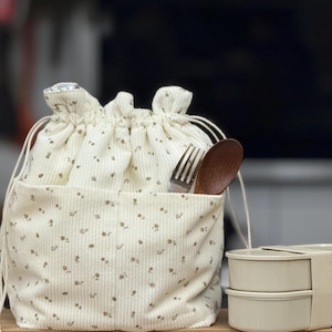 Puede incluir: Bolsa de almuerzo color crema con un estampado floral, cierre de cordón y un bolsillo frontal. Una cuchara de madera y un tenedor están en el bolsillo. Un juego de dos recipientes de comida beige está al lado de la bolsa.