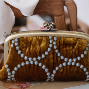 May include: A rectangular mustard-colored velvet coin purse featuring a light blue embroidered circular pattern. The purse has a brushed brass-toned metal frame with a double-knob clasp closure at the top and a small metal loop on the side.