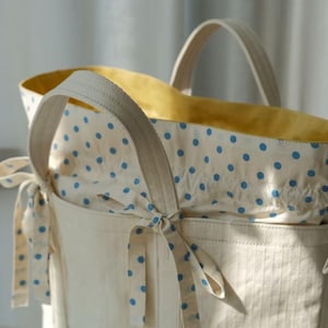 Könnte beinhalten: Cremefarbene Tragetasche mit gelbem Innenfutter und blauem Polka-Dot-Futter. Die Tasche hat zwei Griffe und einen Kordelzugverschluss. Die Außenseite hat ein vertikales Streifenmuster und eine Schleife.