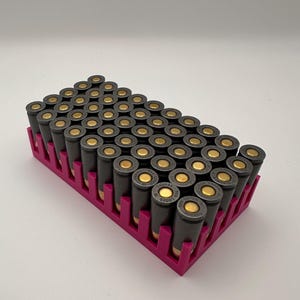 May include: A magenta tray holding numerous dark grey and gold bullet casings. The casings are arranged in rows, highlighting their metallic finish and the contrast with the pink tray. The image is well-lit.
