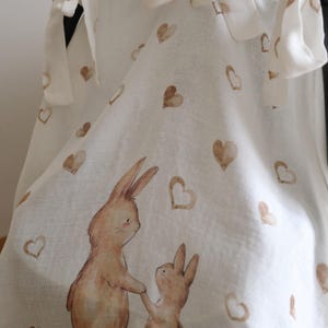May include: White fabric with a repeating pattern of brown hearts and a watercolor illustration of two rabbits holding hands. The fabric is tied with white bows.