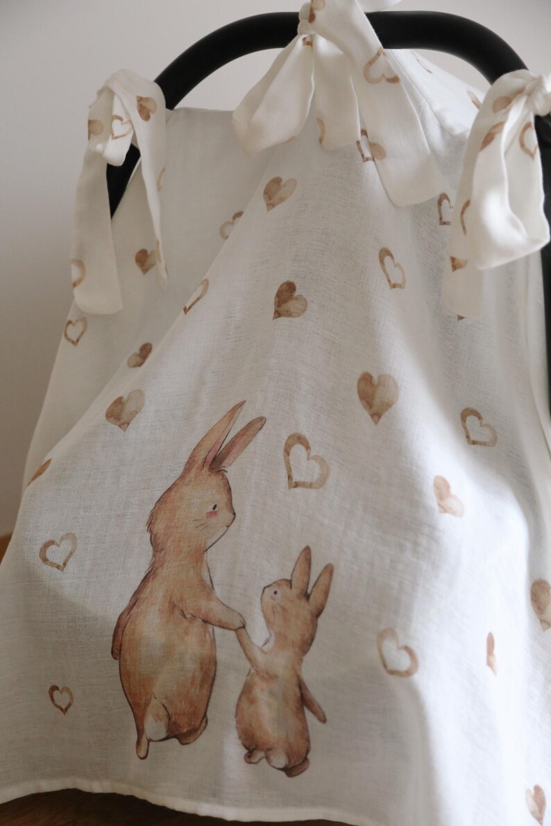 May include: White fabric with a repeating pattern of brown hearts and a watercolor illustration of two rabbits holding hands. The fabric is tied with white bows.