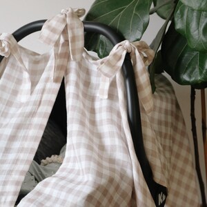 May include: A beige and white gingham patterned car seat cover. The cover is tied to the black car seat handle with matching fabric ties. A large green plant is in the background.