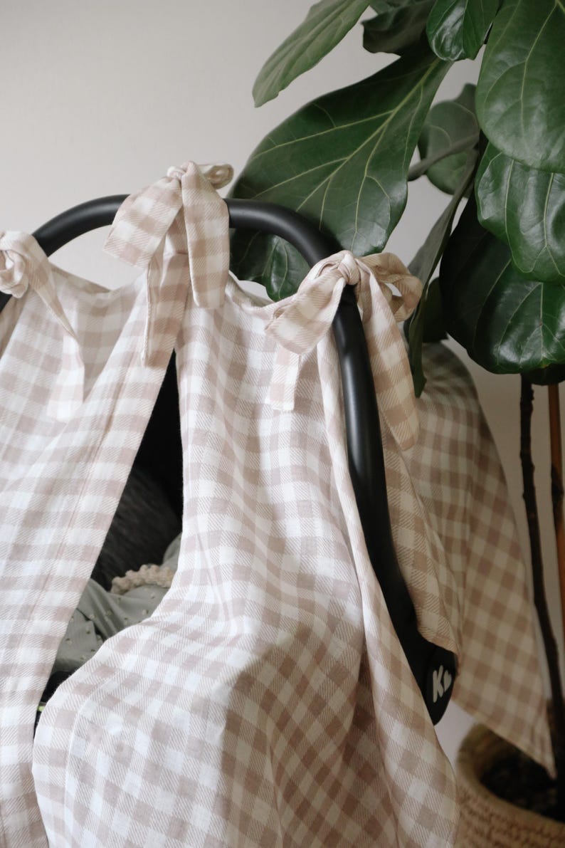 May include: A beige and white gingham patterned car seat cover. The cover is tied to the black car seat handle with matching fabric ties. A large green plant is in the background.