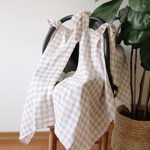 May include: A beige and white gingham patterned car seat cover draped over a black car seat frame. The cover is tied at the top with matching fabric ties. A potted plant is in the background.