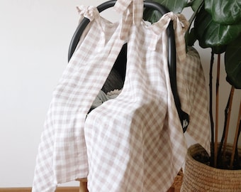 May include: A beige and white gingham patterned car seat cover draped over a black car seat frame. The cover is tied at the top with matching fabric ties. A potted plant is in the background.
