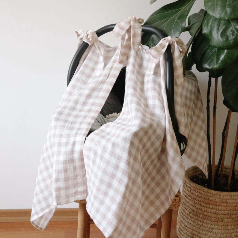 May include: A beige and white gingham patterned car seat cover draped over a black car seat frame. The cover is tied at the top with matching fabric ties. A potted plant is in the background.