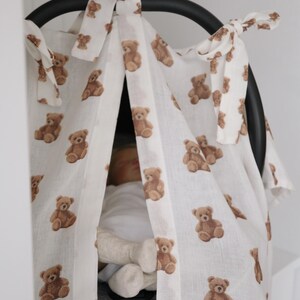 May include: White baby car seat cover with a repeating pattern of brown teddy bears. The cover is tied to the car seat with white fabric ties. The car seat is black.