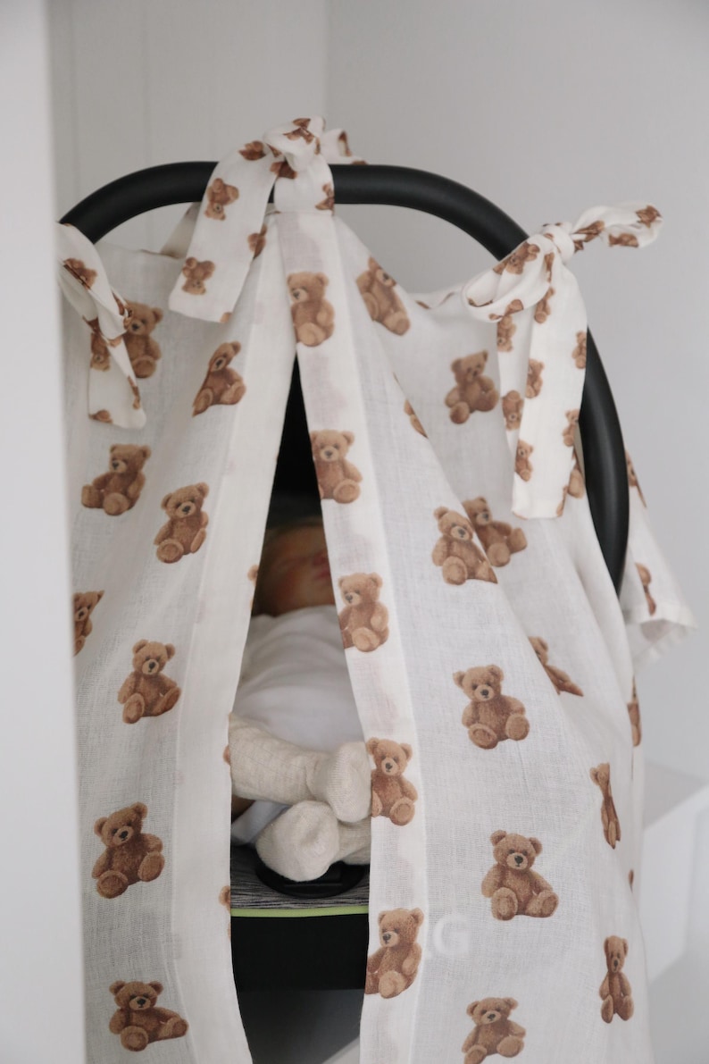 May include: White baby car seat cover with a repeating pattern of brown teddy bears. The cover is tied to the car seat with white fabric ties. The car seat is black.